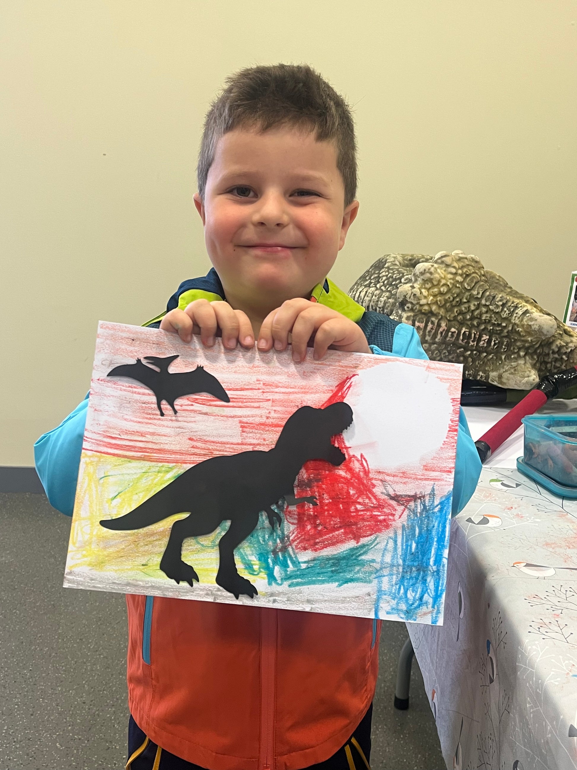 Child holding a drawing of a dinosaur with a pterodactyl on a colorful background.