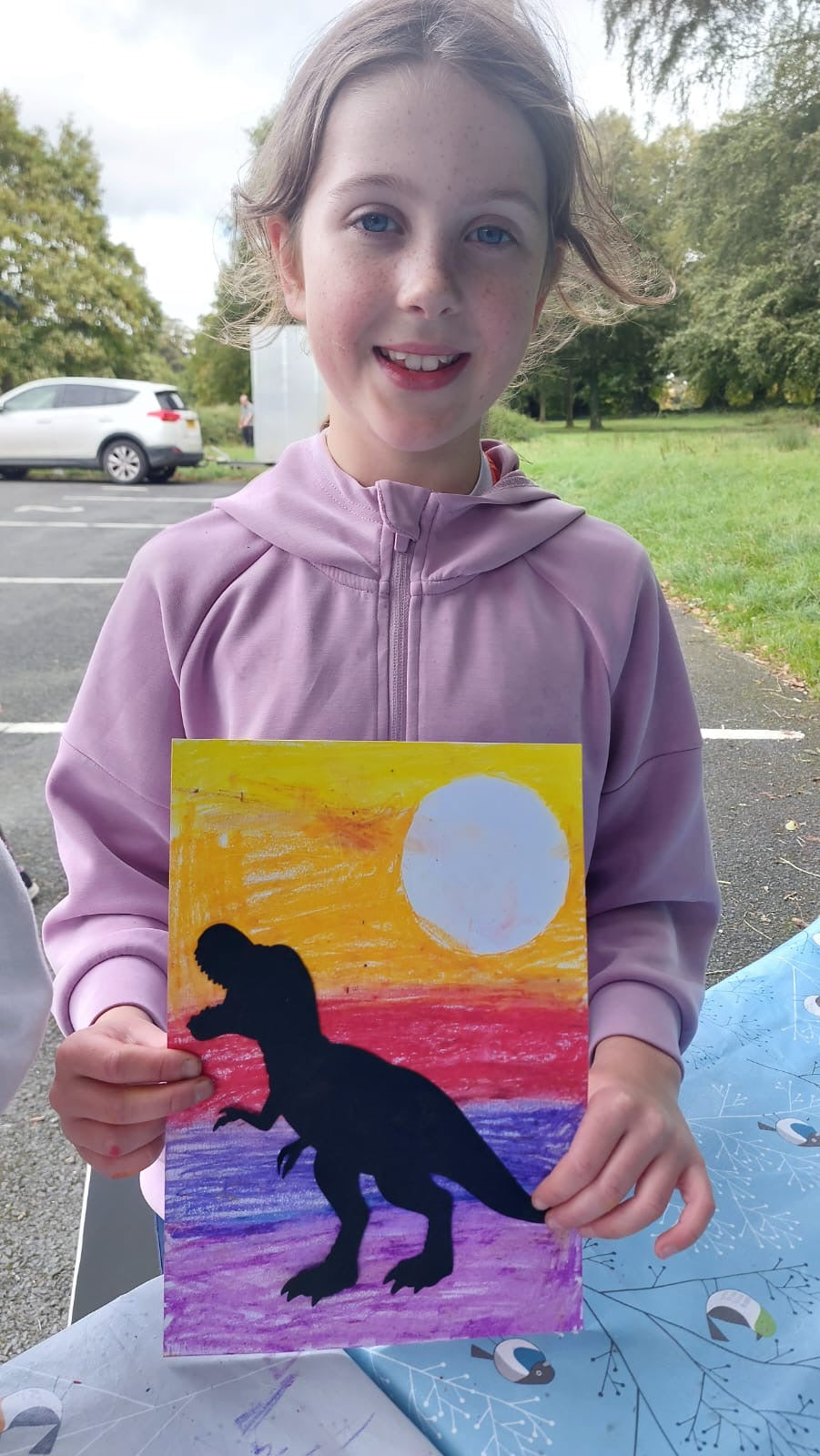 Young girl holding a colourful painting with a dinosaur silhouette outdoors.