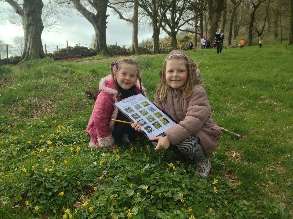 Two children in a park holding a nature activity sheet with trees and grass in the background.