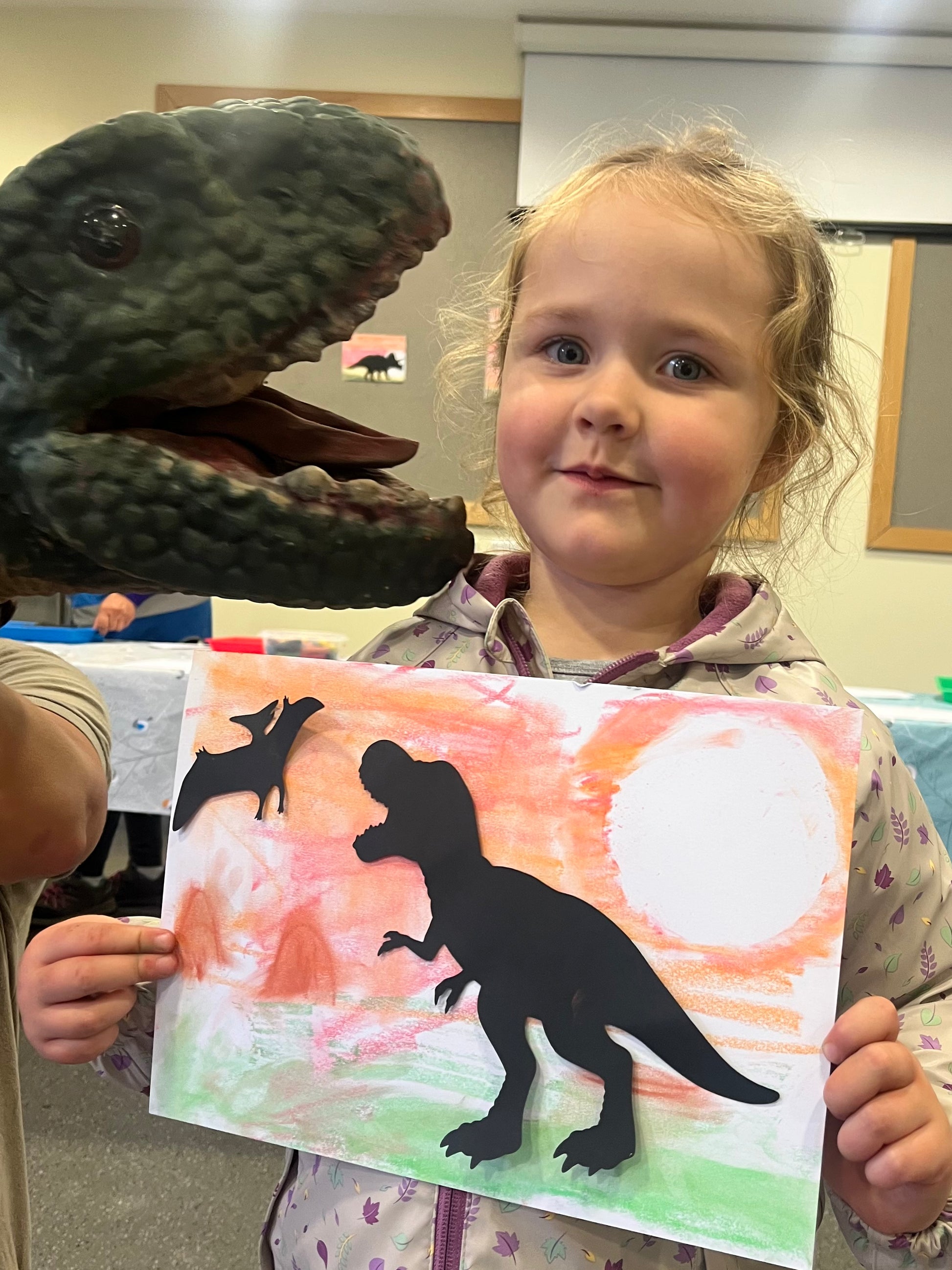 Child holding a dinosaur painting with a dinosaur puppet
