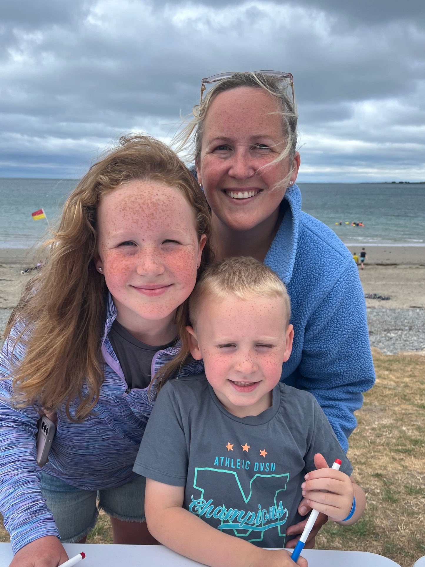 A woman and two children are gathered at a beach table, drawing with markers. The background shows a sandy beach, ocean, and cloudy sky.