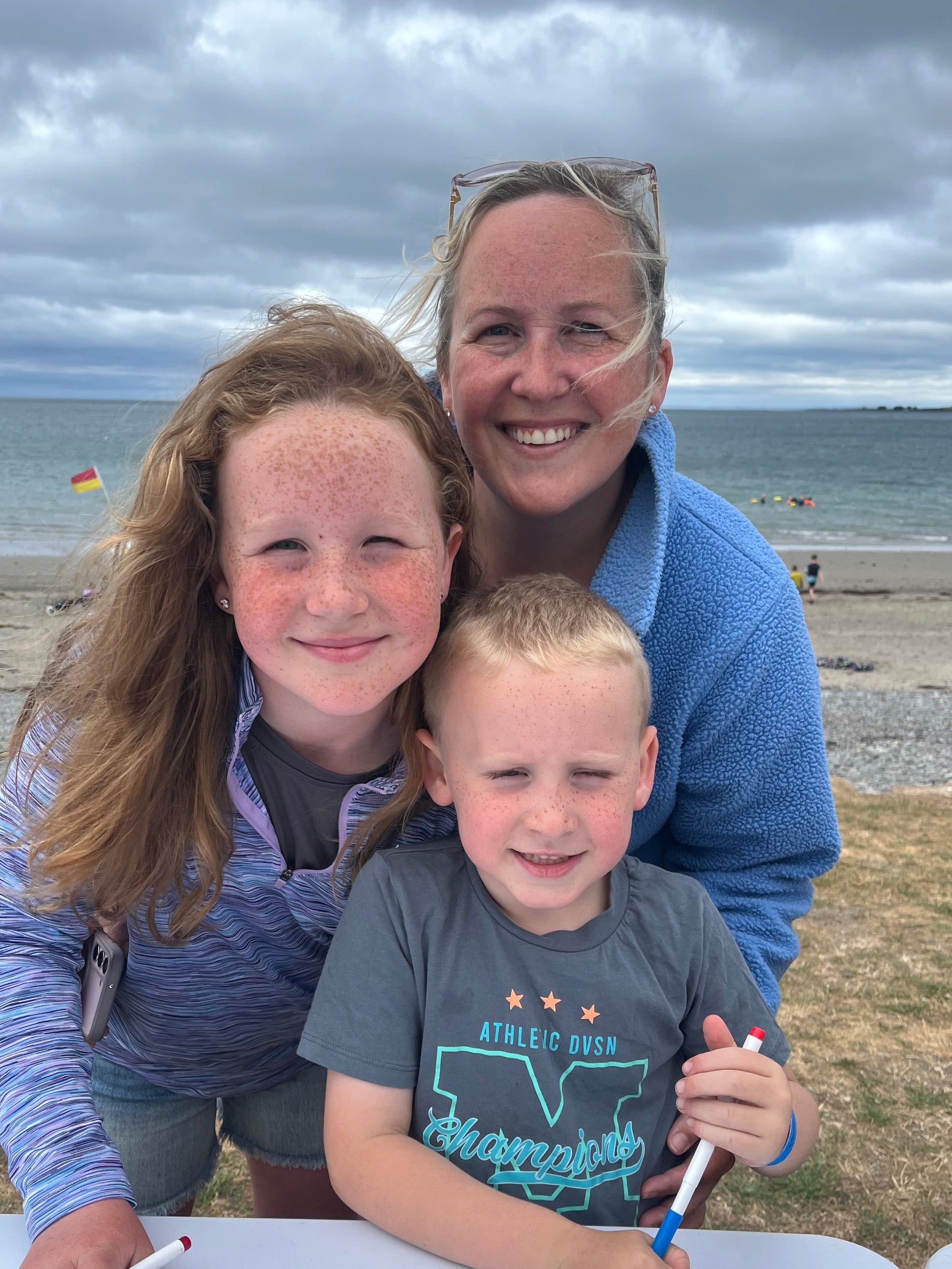 A woman and two children are gathered at a beach table, drawing with markers. The background shows a sandy beach, ocean, and cloudy sky.